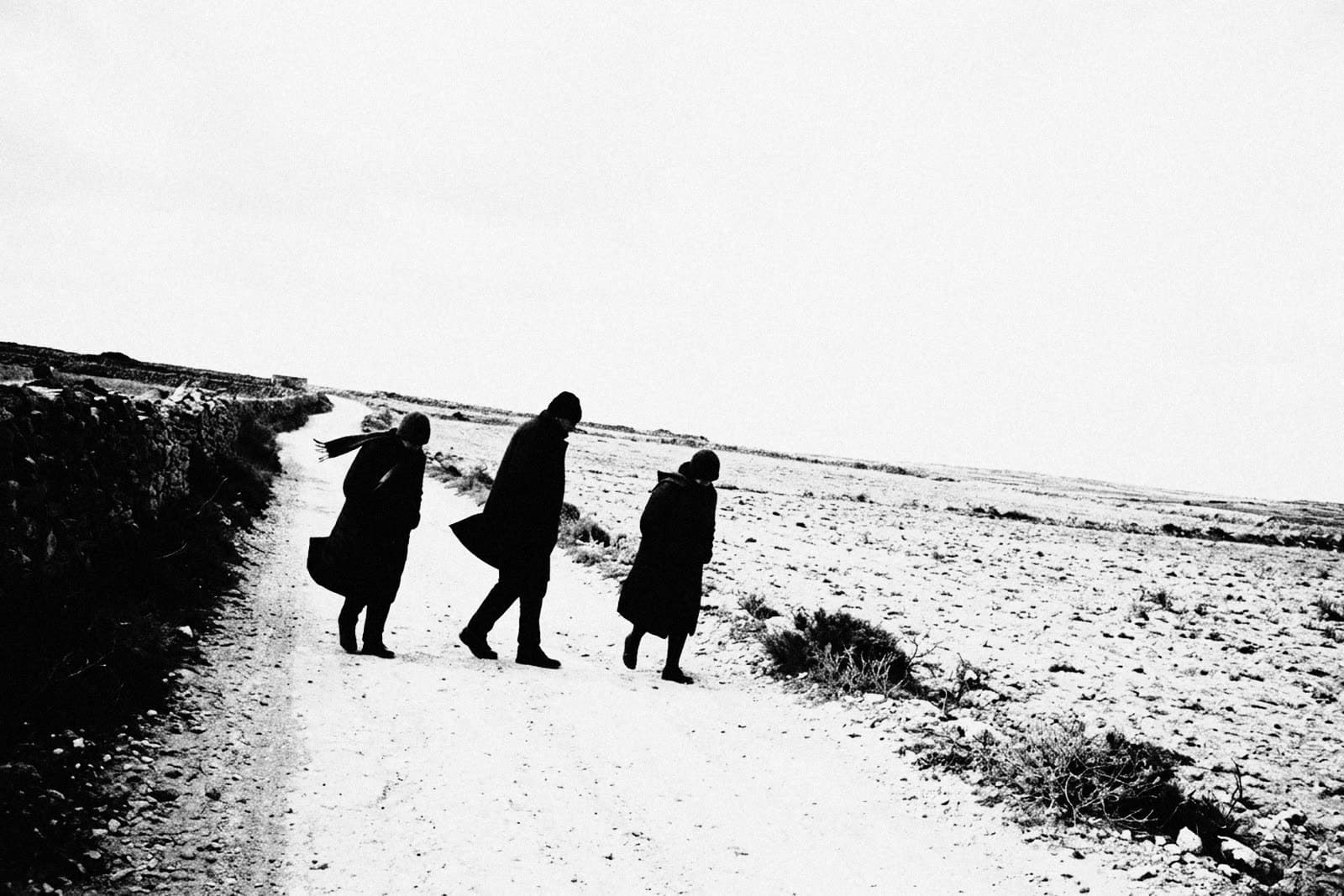 Three people in dark coats walking into the wind on a pale gravel track, coat tails and a scarf blown back, a stone wall on the left, a bare rocky field on the right and a tilted horizon under a white sky