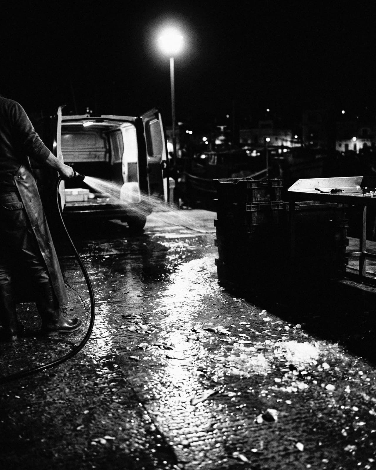 A fish market quay at night, a worker in an apron hosing the wet ground beside an open van