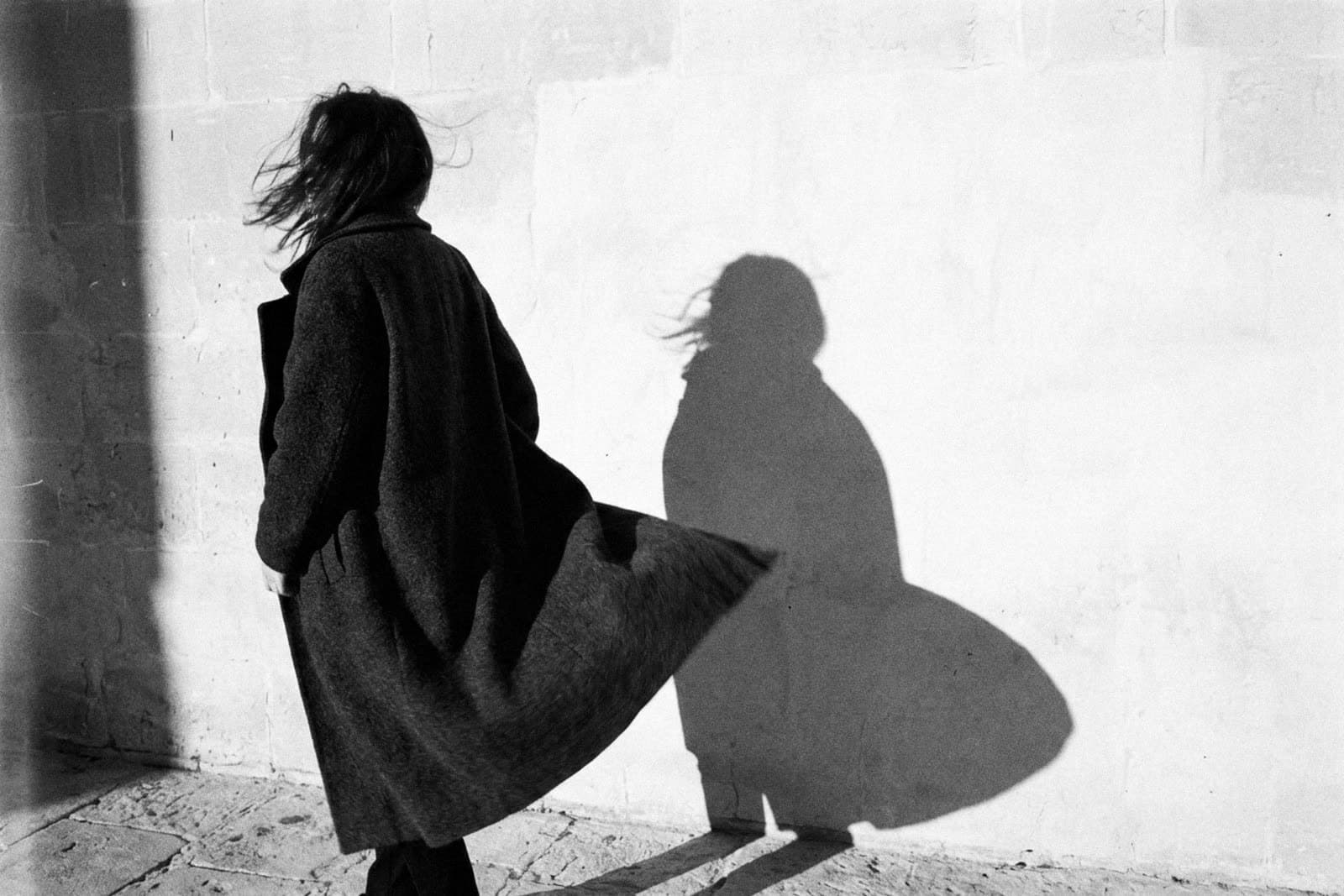 A woman in a long dark coat caught by the wind, her shadow on a pale stone wall