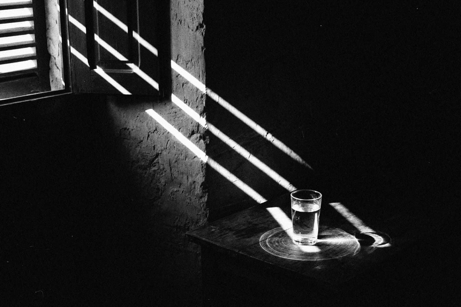 Bars of sunlight from a half-open shutter falling across a rough wall and a small wooden table with one glass of water in a dark room