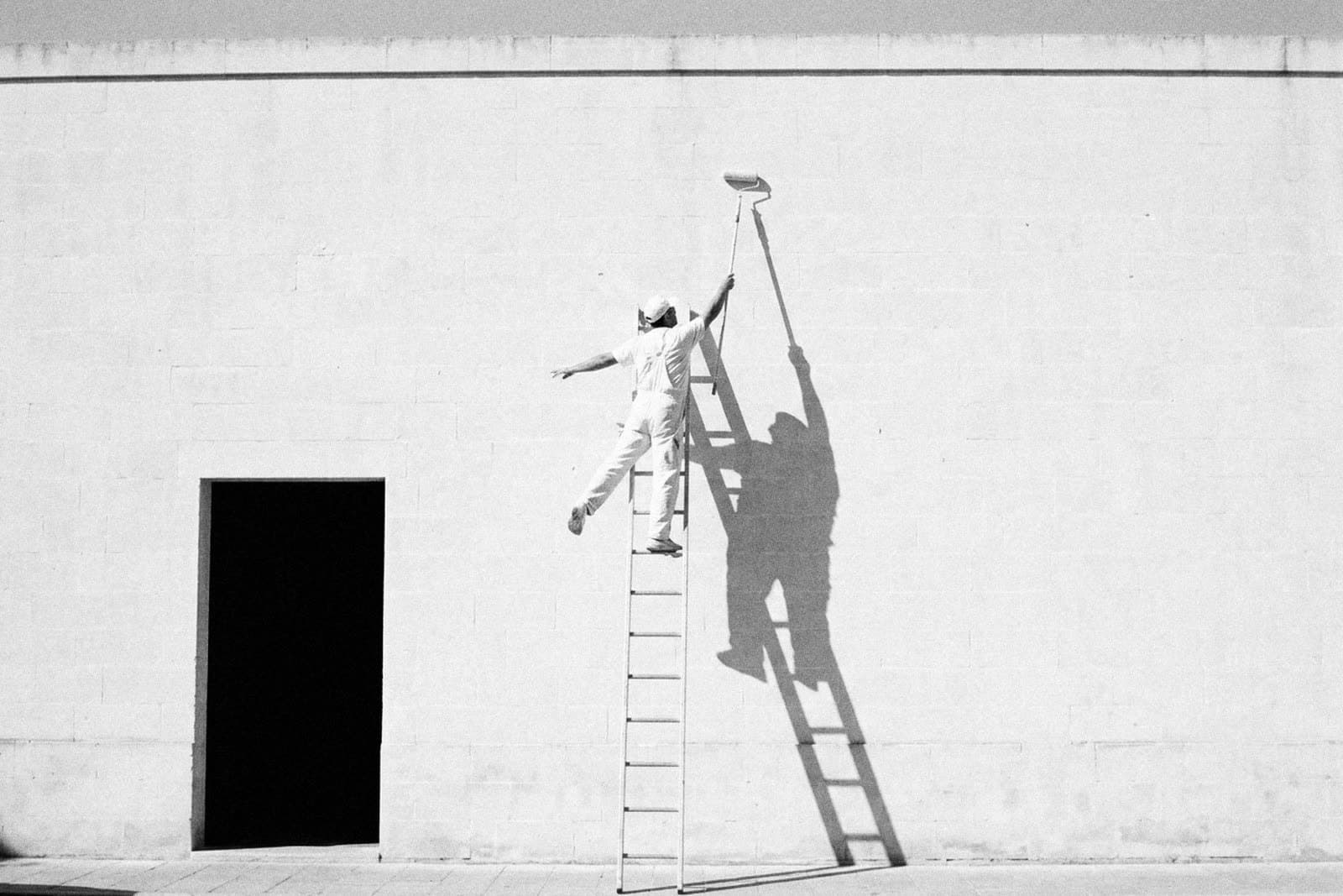 A painter in white overalls and a cap near the top of a tall ladder at a blank limestone wall, one arm stretched up with a roller on a pole, the other out for balance, the long shadow of the man and the ladder on the wall beside him and a black doorway low on the left