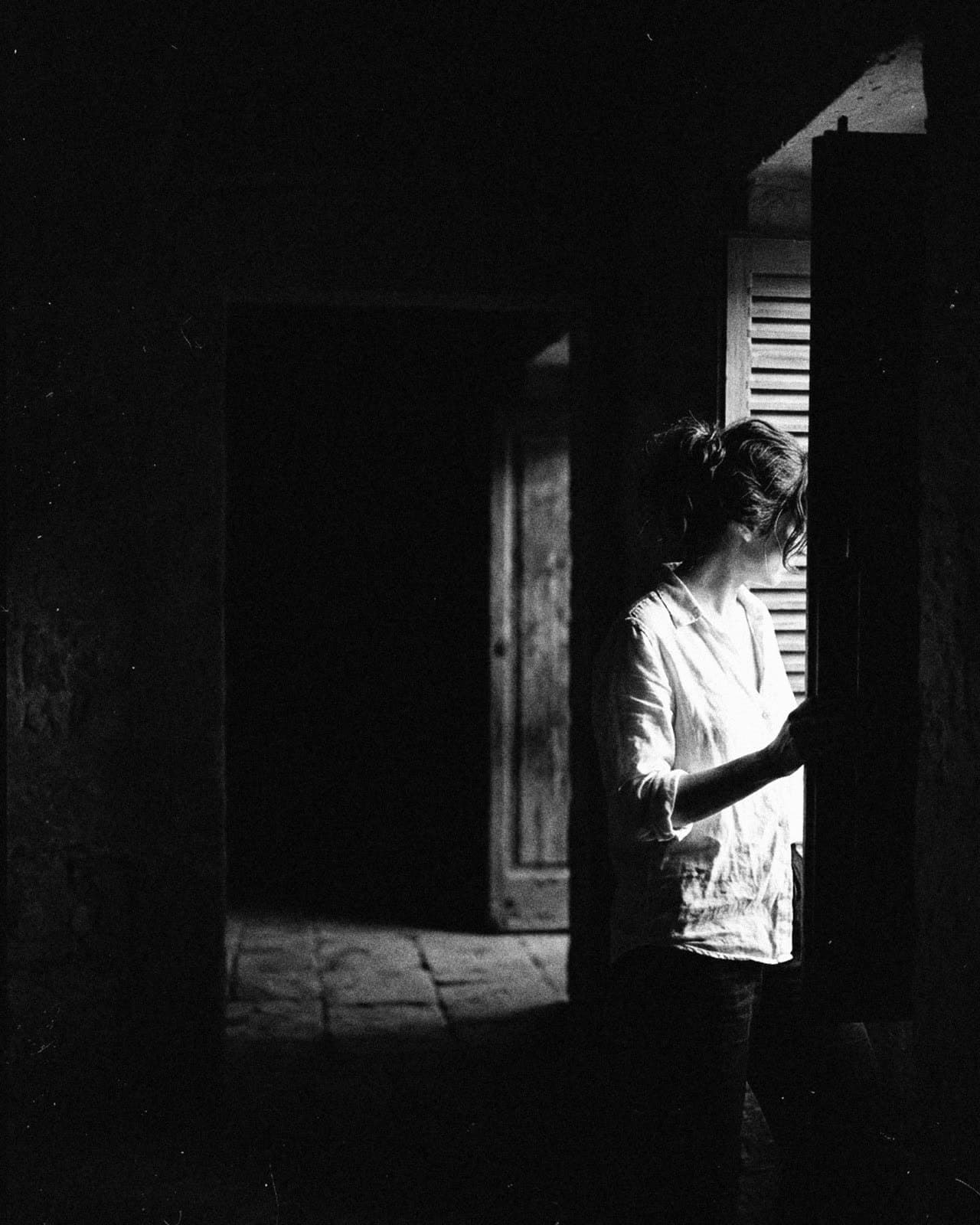 A woman in a white linen shirt by a half-open shutter, the room around her in black
