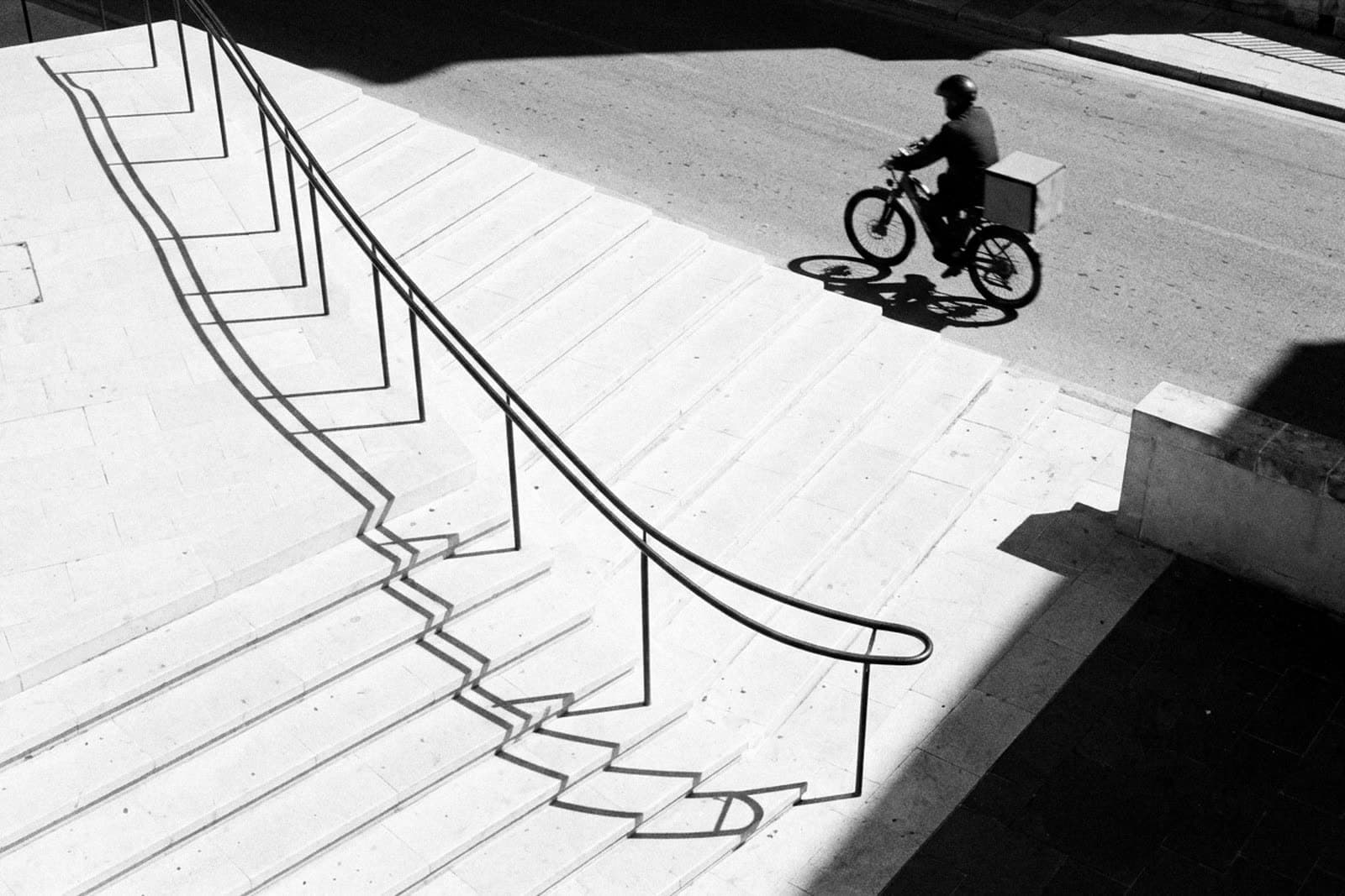 A courier on an e-bike crossing the foot of sunlit stone steps, seen from above, the long shadow of the handrail falling across the steps