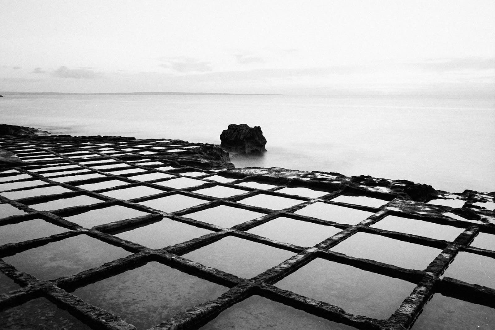 A grid of rock-cut salt pans filled with still water on a flat shore, one dark rock at the edge and a misted sea under a pale sky