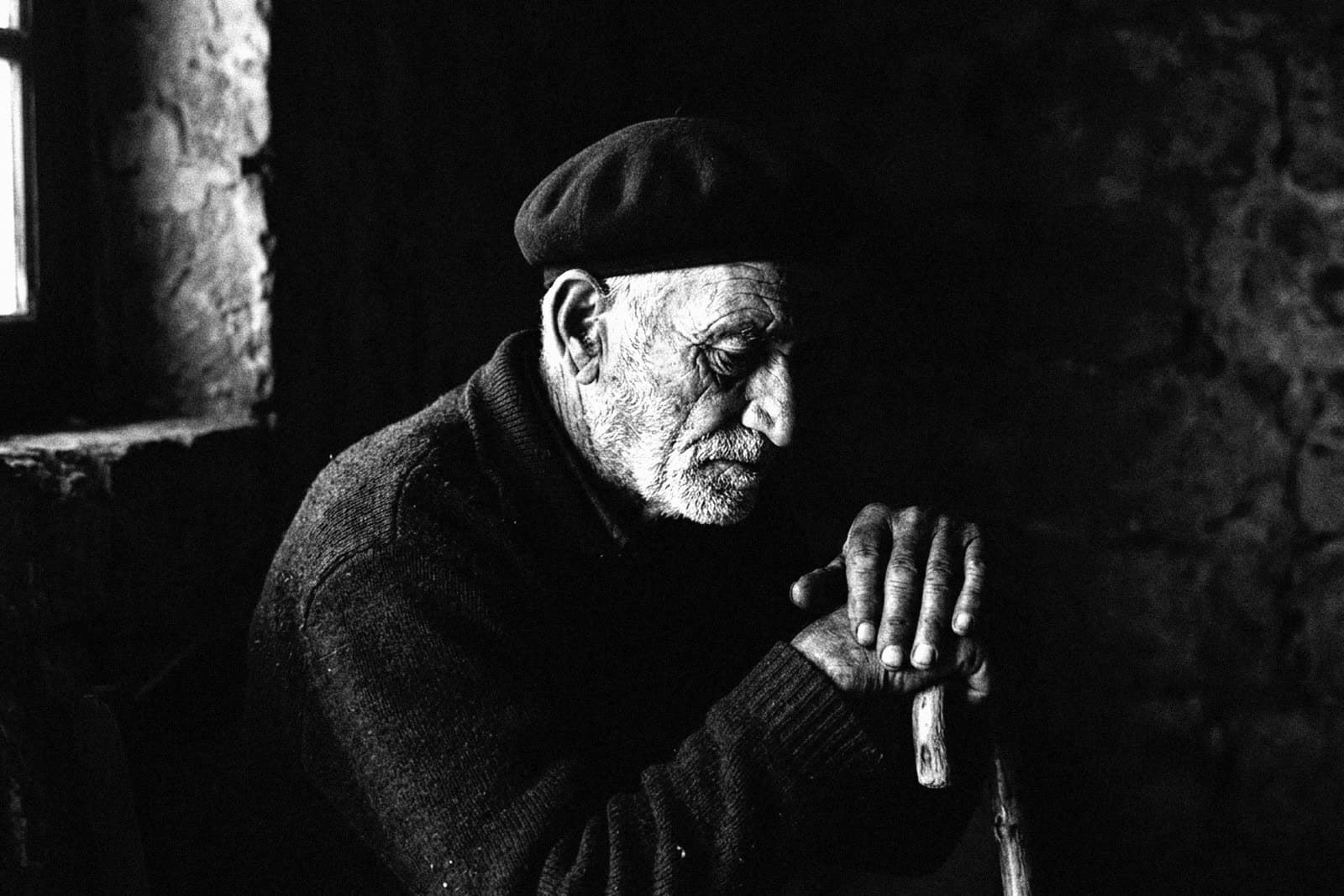 An old man in a dark beret and jumper, in profile with his head lowered, both hands resting on a walking stick, lit by a small window in a dark stone room
