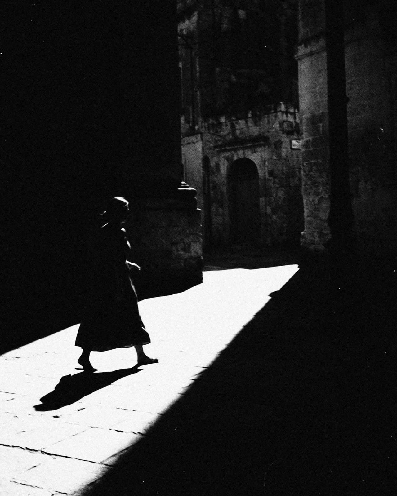 A woman in a dark dress crossing a wedge of hard sun between deep shadows